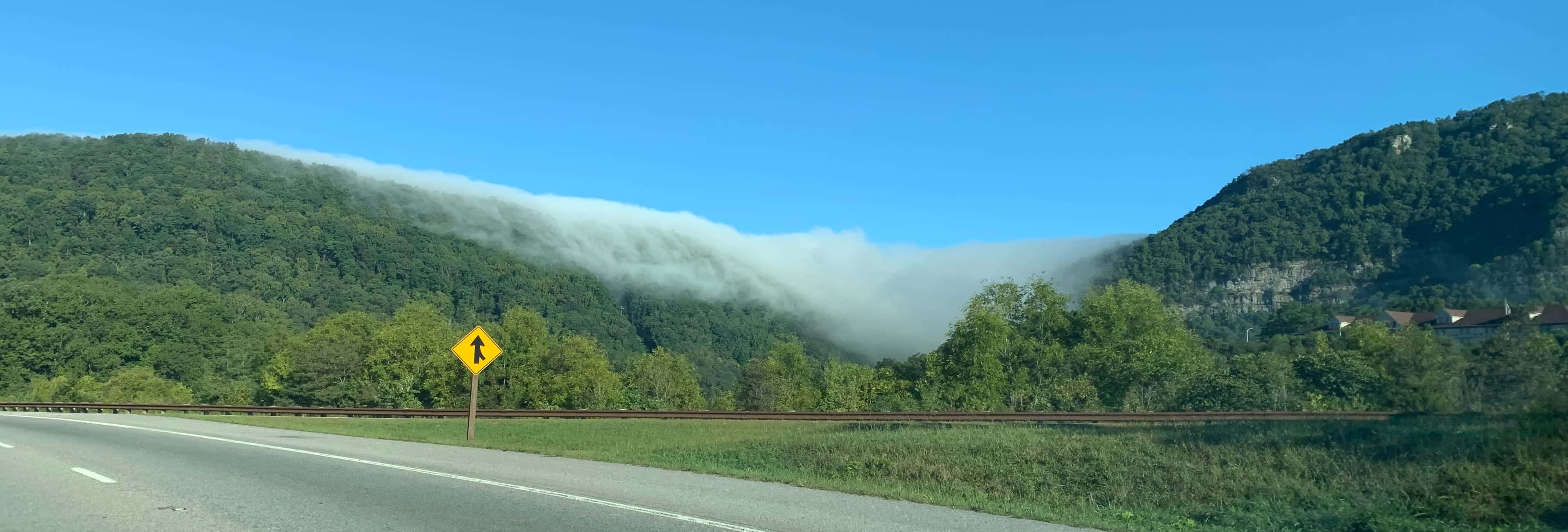 A low cloud spilling over the crest of a forested mountain ridge under a clear blue sky, with a two-lane road and a rail line running along the foot of the slope